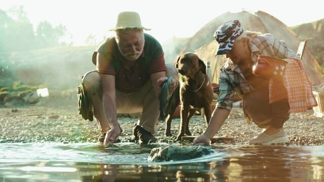 Old man cleaning fresh fish in a stream on a beach, prepares food in nature. A happy retired couple and dog is camping by the river and preparing fish for dinner. Concept of survival in nature