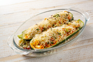 Halved zucchini stuffed with vegetables, feta cheese, parmesan and herbs garnish in a glass baking dish on a wooden table, cooking a healthy vegetarian meal, selected focus