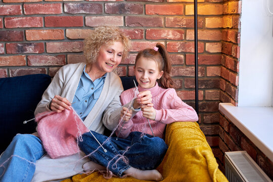 Grandmother And Child Knitting. Senior Woman Teaching Little Girl To Knit. Crafts For Kids. Knitting Tools, Needles. Knitted Hand Made Scarf. Grandmother And Grand Granddaughter Together At Home