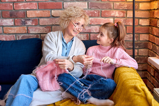 Grandmother And Child Knitting. Senior Woman Teaching Little Girl To Knit. Crafts For Kids. Knitting Tools, Needles. Knitted Hand Made Scarf. Grandmother And Grand Granddaughter Together At Home