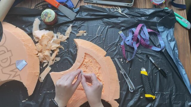Hands cut hatchet form from foam polystyrene with blade on garbage bag. Craftsman's workshop. Woman makes viking axe for cosplay festival. Diy celtic weapon on wooden table.