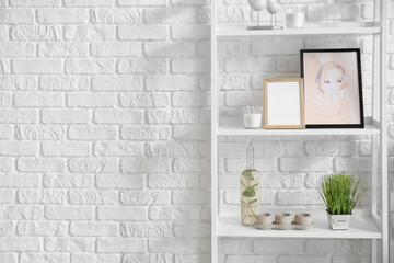 Shelf unit with modern painting, blank photo frame and different decor near white brick wall