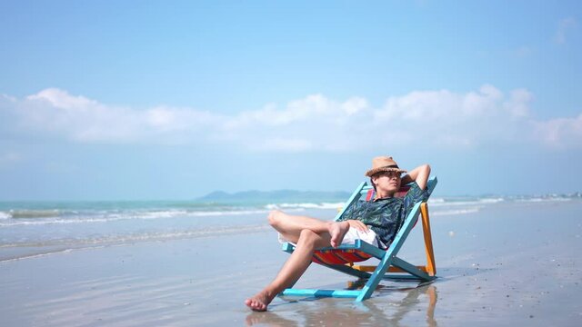Asian man resting on beach chair at tropical beach. Happy guy sunbathing or nap on sunbed by the sea in sunny day. Young man relax and enjoy beach outdoor activity lifestyle on summer holiday vacation
