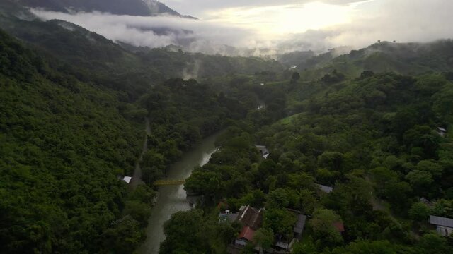 Drone aerial footage of sunset over misty and cloudy rainforest valley and river lined with lush dark green trees near Semuc Champey National Park, Guatemala surrounded by hillsides and mountains.