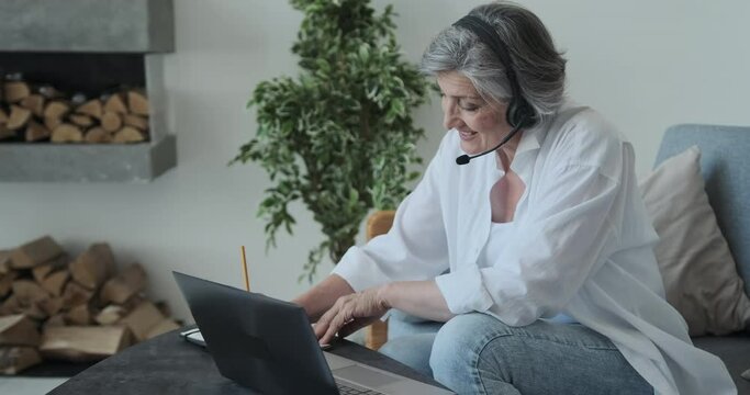 Successful senior mature retired woman lawyer or businesswoman working remotely from a luxury country house, writing notes on a case, using laptop, headset and video call to connect with happy client