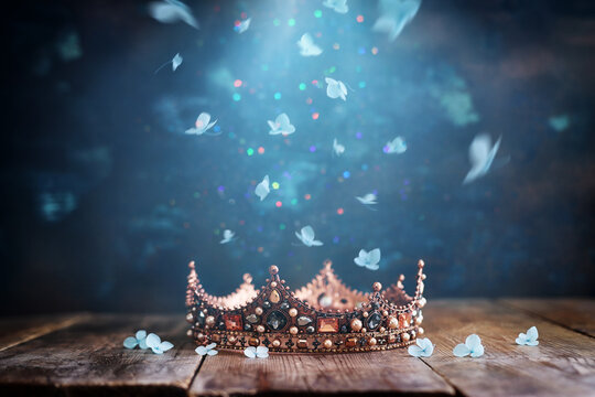 low key image of beautiful queen or king crown over wooden old table and falling flowers. fantasy medieval period
