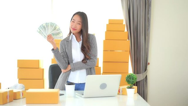 Asian businesswoman working in dropshipping business holding and showing dollar banknotes money in hand standing at her workplace surrounded with yellow packages ready for shipment 