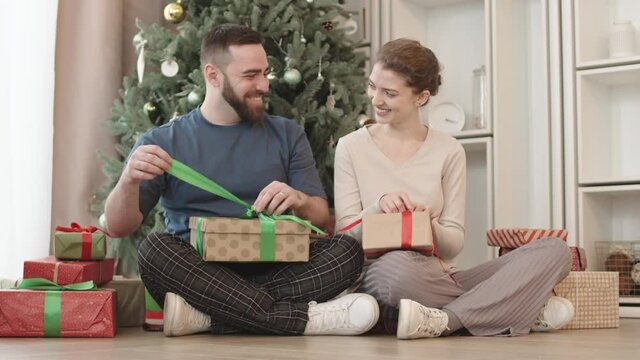 Full shot of happy young Caucasian hetero couple sitting with legs crossed on floor in foreground of Christmas tree at home, smiling and unwrapping gifts in morning