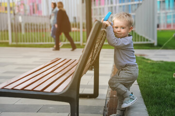 Fototapeta na wymiar Pensive boy with blond hair with a shovel sits on a bench in park on the background of apartment houses and women