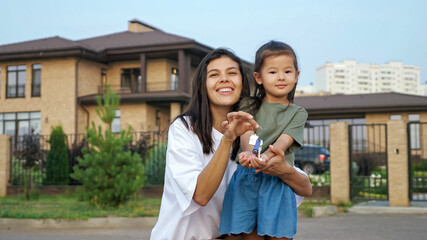 Smiling brunette young woman and little Asian girl boast of keys of new apartment against dwelling buildings on city street closeup