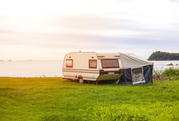 Trailer motor home and a tent on the grassy part of the beach at sunset. Leisure mobile camping home for tourists overlooking the blue sea and cape. Adventure relaxing travel on caravan van - obrazy, fototapety, plakaty