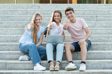 Successful college students gesturing thumbs up or making the ok sign. Happy teenagers sitting on stairs. Teamwork in adolescence concept. - obrazy, fototapety, plakaty