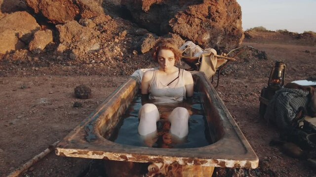 Zoom in shot of young woman looking at camera and submerging under dirty water in old rusty bathtub in post apocalyptic world