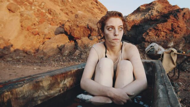 Young shivering woman in wet worn clothes sitting in old dirty bath and looking in distance in post apocalyptic world
