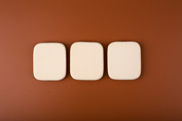 Creative flat lay with three make up sponges isolated on dark brown background. Concept of make up and visage for dark skin tones