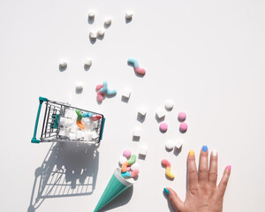 Shopping cart with shugar cubes. Hand and various tasty snacks in pink and mint green, on white background. Excess of sweets, chocolate, candy, sweet food. Sunlight, long shadows.