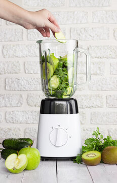 Blender with green fruits and vegetables on a light background. Preparation of a healthy smoothie. The hand puts a piece of apple in the blender bowl.