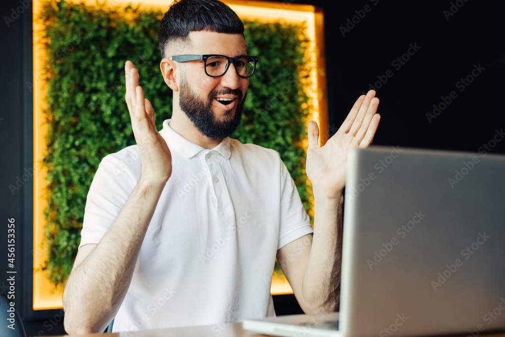 Wall mural portrait of cheerful young man in white t-shirt and business glasses using laptop and having video c