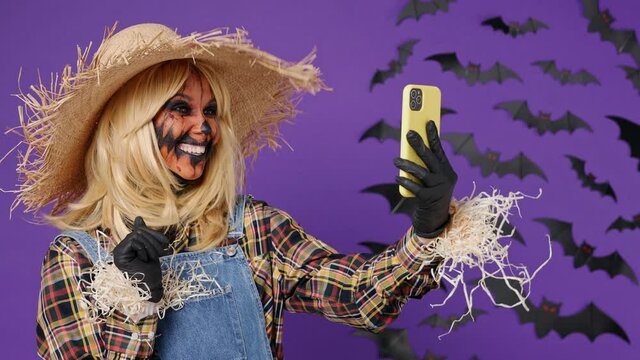 Happy fun young woman with Halloween makeup mask wears straw hat scarecrow costume get video call using mobile cell phone talk greet with hand isolated on plain dark purple background studio portrait