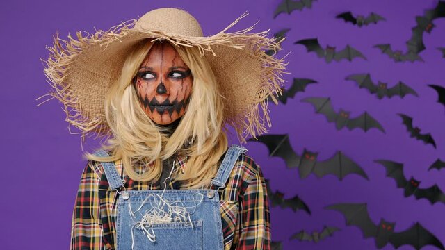 Fun young woman with Halloween makeup mask wears straw hat scarecrow costume looking around raising eyebrows isolated on plain dark purple background studio portrait. Celebration holiday party concept