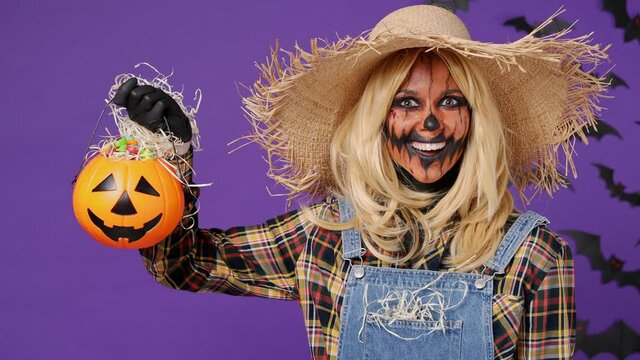 Scary happy smiling vivid young woman with Halloween makeup mask wears straw hat scarecrow costume turn aside take pumpkin basket with candies isolated on plain dark purple background studio portrait