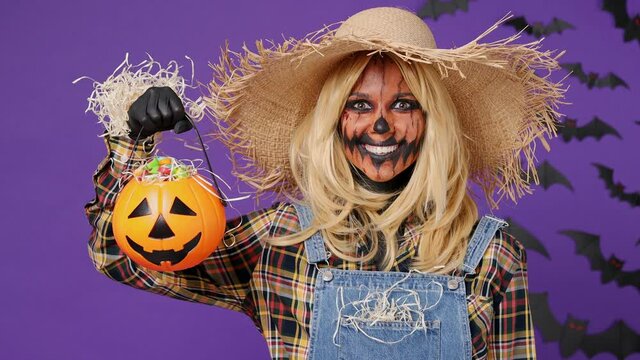 Scary happy smiling young woman with Halloween makeup mask wears straw hat scarecrow costume raise pumpkin basket with candies nodding say yes isolated on plain dark purple background studio portrait