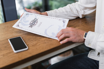 cropped view of man holding menu near smartphone with blank screen on table