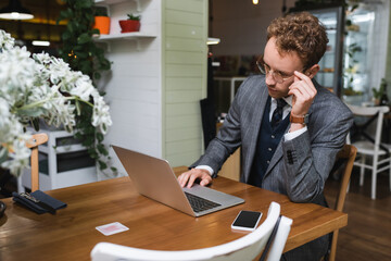 curly businessman in suit adjusting glasses while using laptop in cafe