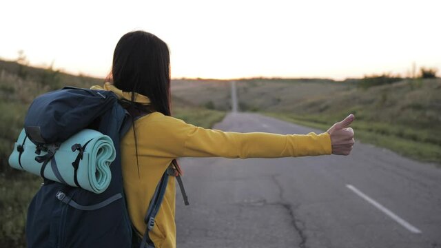 The concept of traveling and hitchhiking. A young female traveler hitchhiking and stopping car with thumbs up on road. A female hitchhiker by the road during vacation trip. Summer vacation adventure.