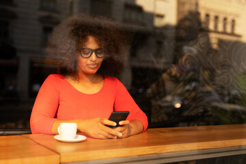 Young woman at cafe drinking coffee and using mobile phone. Beautiful African woman typing a message.
