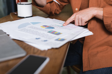 cropped view of young man pointing at charts and graphs near gadgets in cafe
