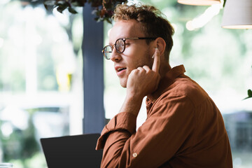 young freelancer in glasses adjusting earphones near laptop with blank screen in cafe