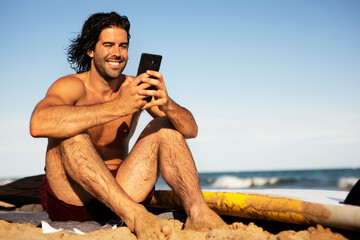 Portrait of handsome surfer with his surfboard. Young man using the phone while relaxing on the beach.