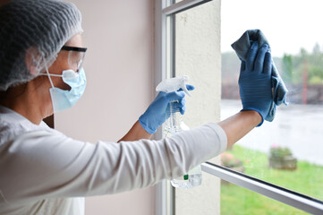 Naklejka premium Mature woman in protective medical suit and respirator mask desinfecting furniture and cleaning window glass at hospital