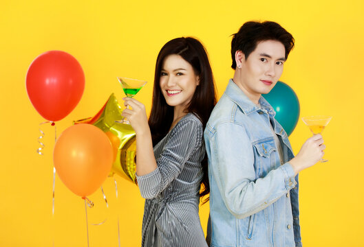 Studio shot of Asian happy gay and female friend couple in fashionable jeans wears standing look at camera lean on each other back holding cocktail glasses in celebration party on yellow background