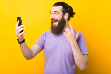 Excited bearded man is using his earphones and phone over yellow background.