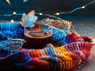 Autumn coffee on a dark table with a bright multicolored knitted warm scarf, a garland and maple...