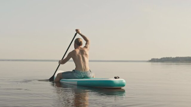 Slowmo rear-view shot of young athletic man sitting on sup paddle board paddling in lake at sunset