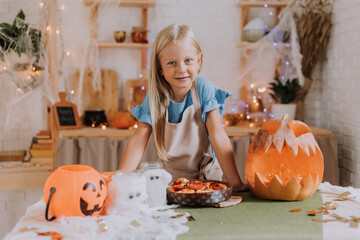 a blonde baby girl with long hair in an apron in the kitchen, decorated with pumpkins and garlands for Halloween, prepares a focaccia pie. space for text. High quality photo