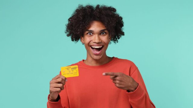 Happy young curly african american man 20s wears azure t-shirt pointing index finger on mockup plastic credit bank card showing thumbs up isolated on plain pastel light blue background studio portrait