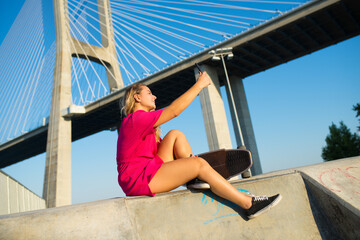 Pretty young woman taking selfie. Beautiful girl in crimson dress with skateboard sitting near bridge, taking photo of herself. Sport, hobby, active lifestyle, social media, photo concept
