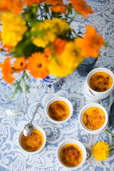 French dessert creme brulee with caramel crust in white baking molds on blue and white tablecloth and a bouquet of yellow and red marigolds flowers
