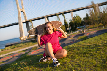 Naklejka premium Portrait of attractive young woman with longboard sitting on grass. Beautiful girl in crimson dress with skateboard behind back sitting at park. Sport, hobby, active lifestyle concept