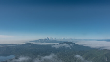 Kamchatka landscape, view from a height. Clouds float over the green mountains and the lake. In the distance, against the blue sky, a ridge of conical volcanoes with snow-covered slopes