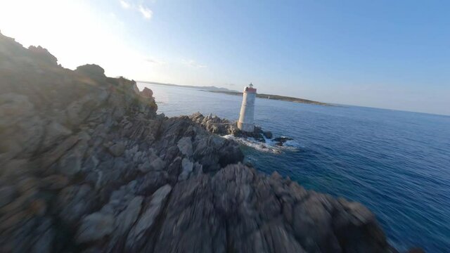 FPV video, view from above, stunning aerial view from an FPV drone flying at high speed over a rocky coastline with a lighthouse illuminated during a dramatic sunset. Faro di Capo Ferro, Sardinia.