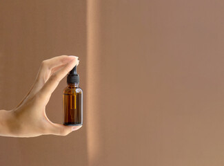 Mockup of dropper bottle is located between two fingers of woman's hand on brown background. Close-up, copy space. Concept of body care, cosmetology