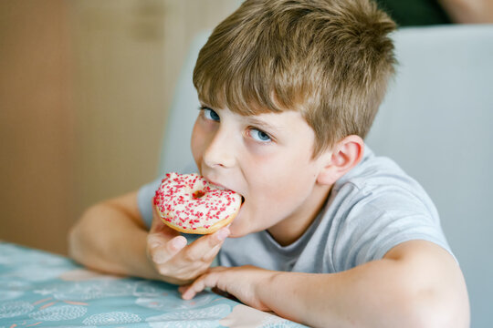 Happy preteen school boy eating sweet donut indoor. Blond child in domestic kitchen or in school canteen Cute kid and unhealthy sugared food.