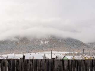 The village is covered in snow, the roofs of houses can be seen behind the fence. Altai mountains are covered with thick clouds. Chemal, Altai Republic, Russia.