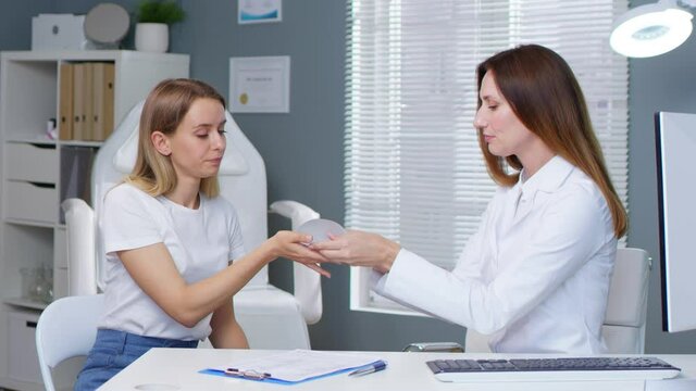 Plastic surgeon showing breast implant to her female patient. Blonde young woman choosing breast implants before breast augmentation surgery. Beauty clinic concept