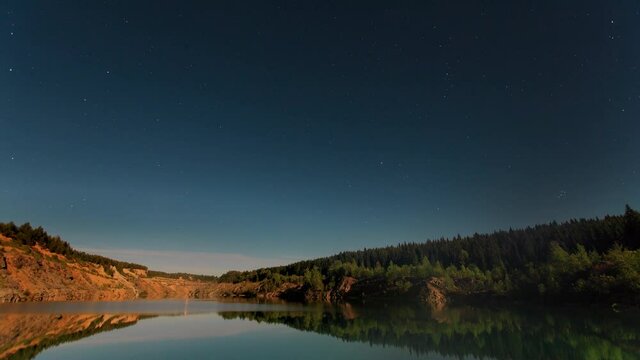 Timelapse landscape with a lake, in the sky above the lake there are stars. Summer lake next to a cliff against the backdrop of a starry evening sky.